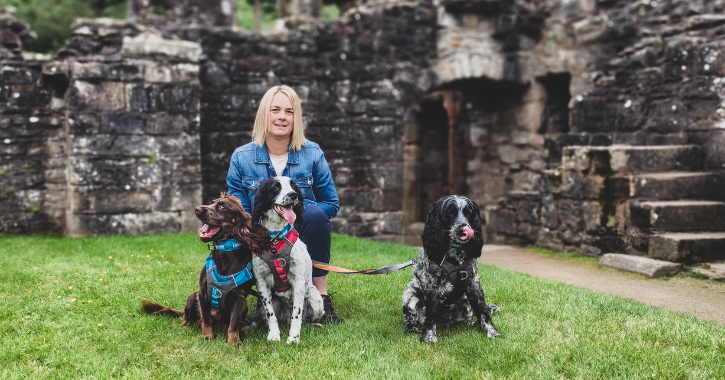 Woman sat with three dogs in the grounds of Finchale Priory, with the priory ruins visible in background.